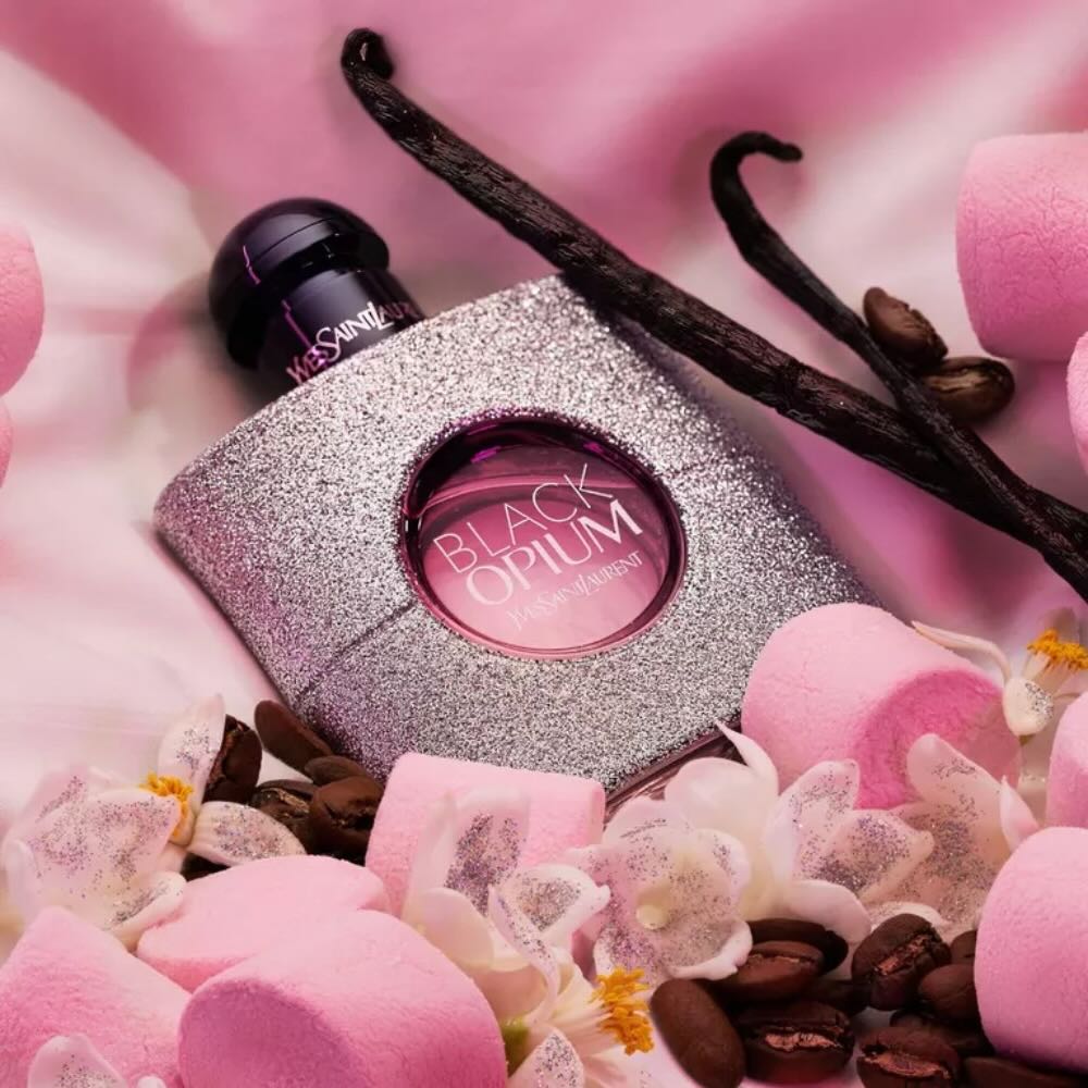 YSL Black Opium perfume bottle surrounded by vanilla beans, coffee beans, and pink flowers on a pink background