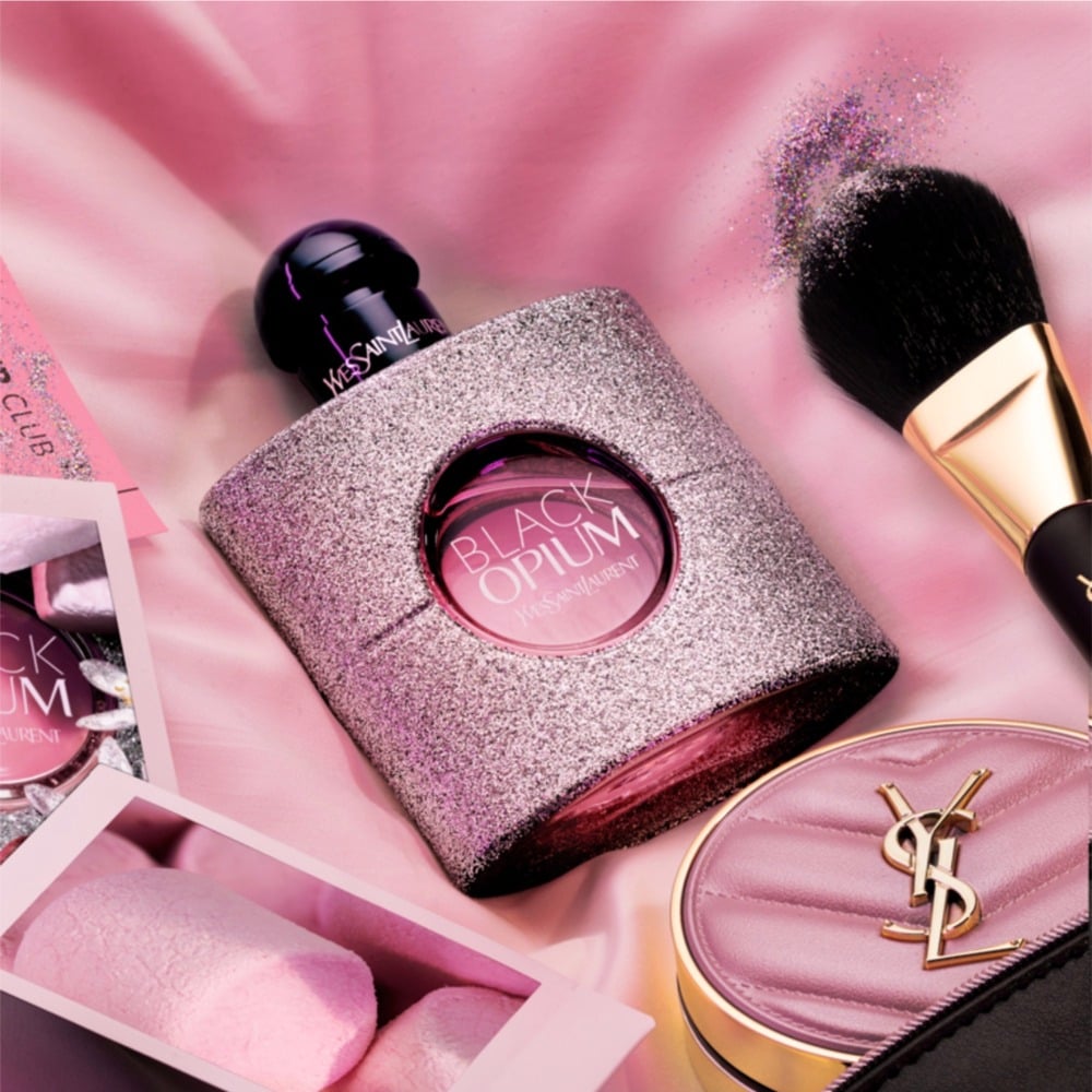 YSL Black Opium perfume bottle with makeup items on a pink background