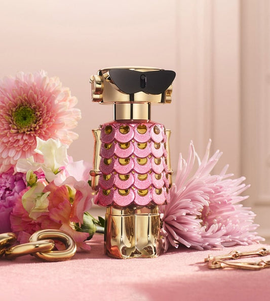Perfume bottle with pink and gold design surrounded by flowers on a pink background Perfume bottle with decorative cap on a pink background with flowers Paco Rabbane Fame Blooming Pink 80ml