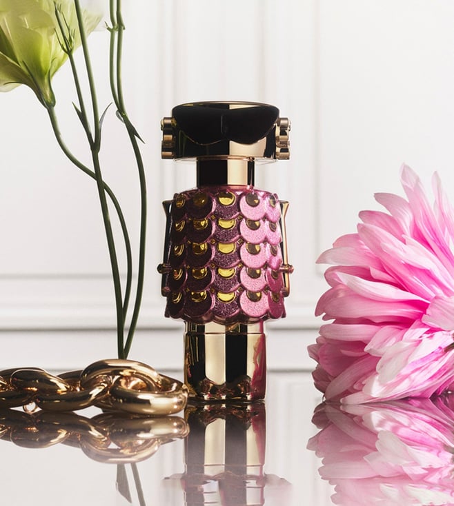 Perfume bottle with decorative casing next to a pink flower on a reflective surface Perfume bottle with decorative cap on a pink background with flowers Paco Rabbane Fame Blooming Pink 80ml
