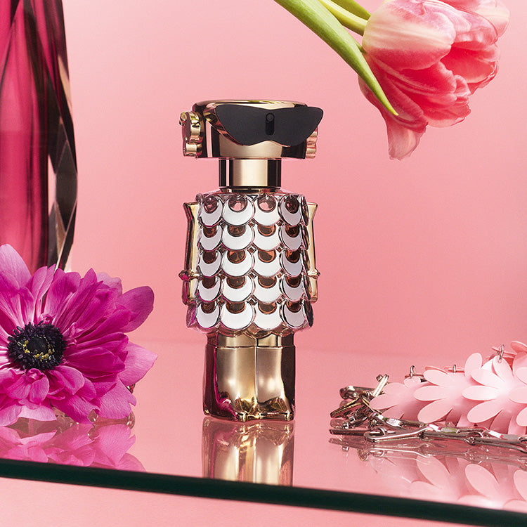 Perfume bottle with decorative cap on a pink background with flowers Paco Rabbane Fame Blooming Pink 80ml