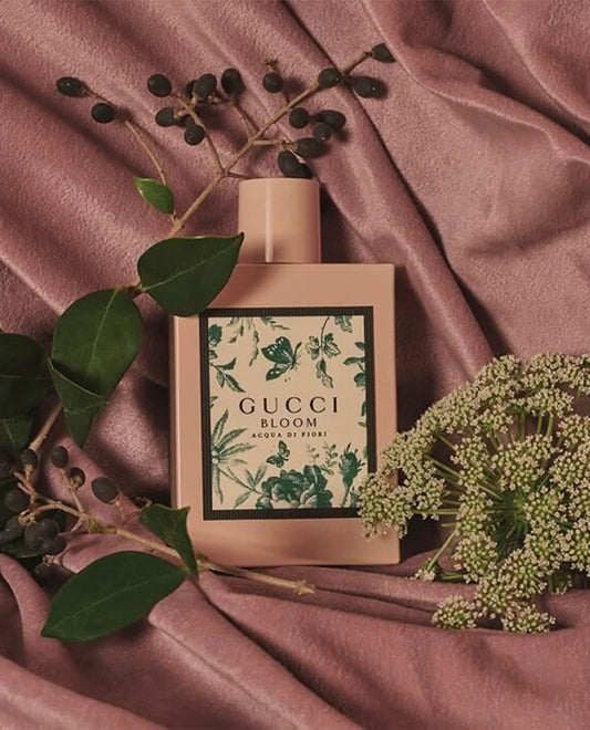 Gucci Bloom perfume bottle on a pink fabric background with green leaves and flowers. 