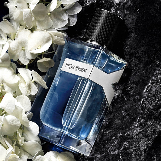 Blue Yves Saint Laurent perfume bottle on a dark surface with white flowers. Yves Saint Laurent Y Eau de Toilette 100 ml for Men With Fresh Woody Masculine Fragrance
